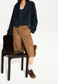 Laivastonsininen lyhyempi takki, vaaleat cargo-shortsit, ruskeat nauhaliuku kengät paksuilla pohjilla ja valkoiset nilkkasukat, poseerattuna tummalla tuolilla vaaleaa taustaa vasten.