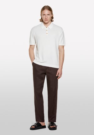 Polo shirt blanc à manches courtes avec un tissu texturé et trois boutons orange, associé à un pantalon en lin brun foncé et des sandales noires.