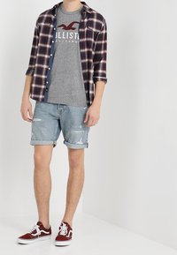 Rutiga skjortor i marinblått, rött och vitt över en grå grafisk T-shirt; slitna denimshorts; röda och vita sneakers. Avslappnad outfit.