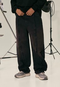 Black denim pants with frayed side seams and a relaxed fit. Paired with grey sneakers, set against a neutral background.