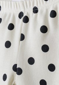 Bílá ribbová tkanina s velkými černými puntíky po celém povrchu. Textura elastického pasu je viditelná, což zdůrazňuje neformální design.
