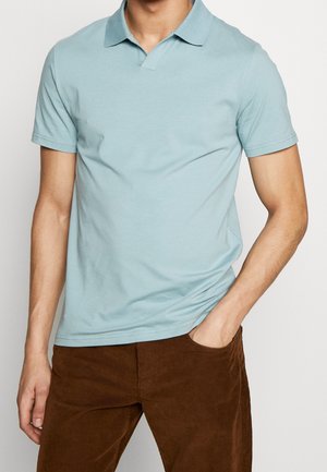 Homme portant un polo bleu clair à manches courtes avec un pantalon en velours côtelé marron, la main posée dans la poche, sur un fond uni.