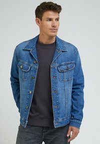 Mężczyzna w niebieskiej dżinsowej kurtce na ciemnoszarym swetrze z okrągłym dekoltem, stojący z rękami w kieszeniach, lekko zwrócony w prawo.