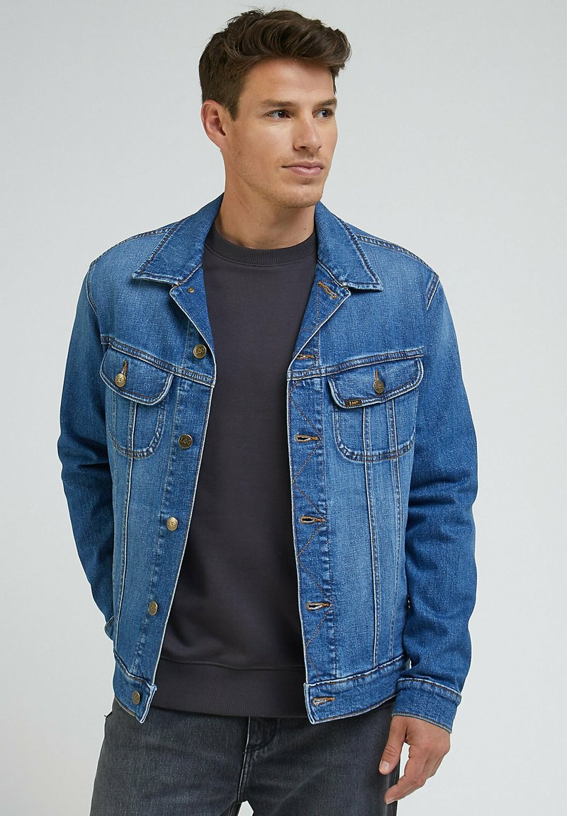 Mężczyzna w niebieskiej dżinsowej kurtce na ciemnoszarym swetrze z okrągłym dekoltem, stojący z rękami w kieszeniach, lekko zwrócony w prawo.