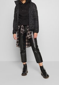 Giacca a vento nera con cappuccio, felpa nera, camicia di flanella a quadri annodata in vita, pantaloni neri lucidi e testurizzati, e stivali neri con lacci.