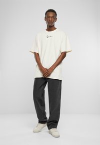 Avslappet passform, kremfarget t-skjorte med korte ermer og svart logo, kombinert med løse, mørk grå jeans og kremfargede sneakers. Glatt bomullstekstur.