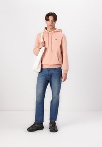 Sudadera rosa claro con un bolsillo frontal y un pequeño logo, combinada con jeans azules y zapatillas negras. El modelo sostiene una bolsa de tela blanca.