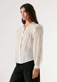 Balta, lengva bluzelė su raukšlėmis ant pečių, klostuota priekine dalimi ir mygtukų uždarymu. Ilgos rankovės su apvadu; šiek tiek ilgesnė nugara.