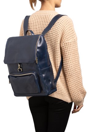 Persoon draagt een beige gebreide trui en zwarte broek, met een marineblauwe leren rugzak met een voorvak en klepsluiting.