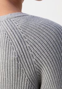 Gros plan sur une personne portant un pull en tricot gris clair, mettant en évidence les détails de la texture des côtes et du point de semence sur l'épaule arrière.