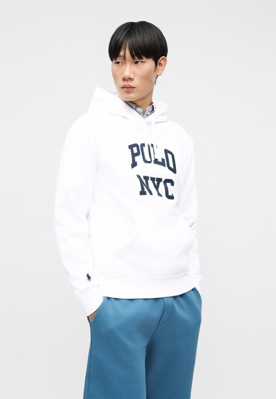 Νεαρός άνδρας που φοράει λευκό φούτερ με κουκούλα "POLO NYC" και μπλε παντελόνι, με τα χέρια στις τσέπες, κοιτάζοντας προς τα πλάγια σε ένα απλό φωτεινό φόντο.