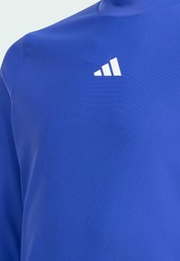 Modrá atletická halenka vyrobená z hladkého, elastického materiálu. Obsahuje vysoký límec a bílé logo se třemi pruhy na levém rameni.