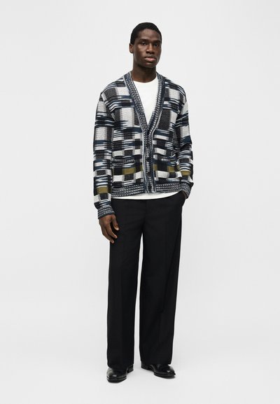 Mężczyzna w czarno-biało-oliwkowym kardiganie w wzory, białej koszuli, czarnych szerokich spodniach i czarnych butach, stojący na jednolitym tle.