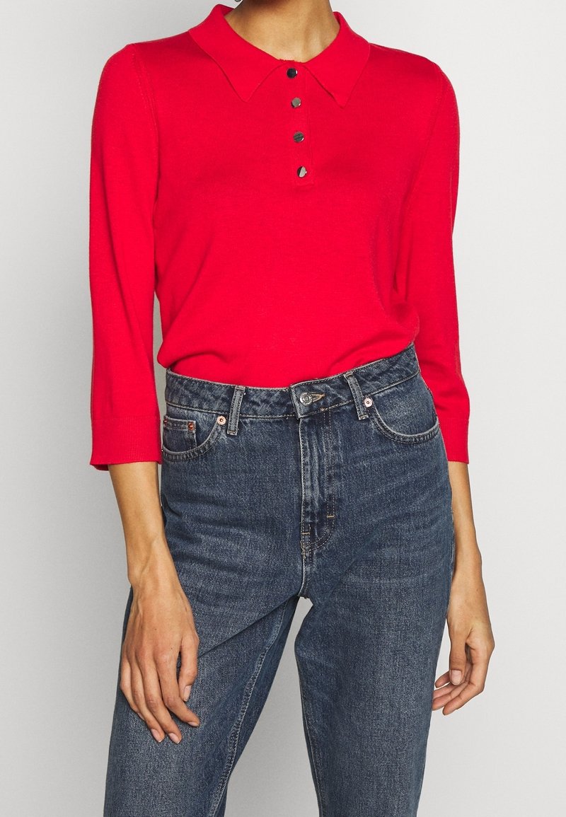 Rotes Langarm-Polohemd mit Kragen und drei Knöpfen, kombiniert mit hochgeschnittenen dunkelblauen Jeans. Glatte Textur, lässige Passform.