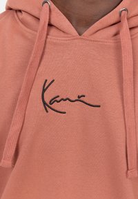 Sudadera con capucha en suave mezcla de algodón en color coral, que presenta un logotipo negro bordado en el pecho y detalles de cordón.