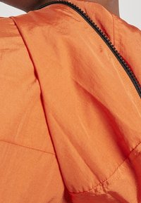 Orange lättviktsmaterial med en slät yta och matt finish, med en svart dragkedja längs sömmen för stängning och detaljering.