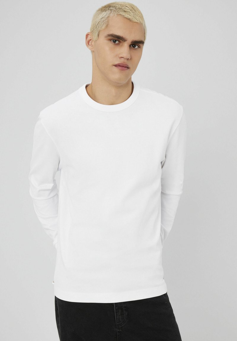 T-shirt blanc à manches longues avec un col rond, texture lisse et silhouette ajustée, associé à un jean foncé.