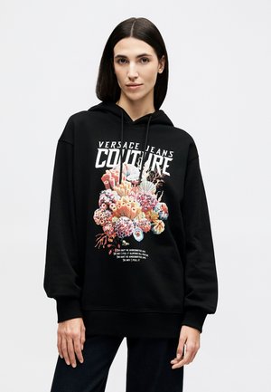 Žena s rovnými tmavými vlasy nosí černou mikinu Versace s barevným motivem korálového útesu a bílým nápisem "Versace Jeans Couture".