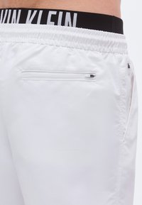 Λευκά αθλητικά σορτς με ελαστική μέση και μαύρο λογότυπο. Διαθέτουν πλαϊνή τσέπη με φερμουάρ και λείο, ελαφρύ ύφασμα.