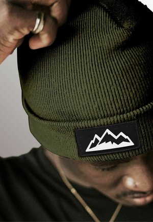 Knit green beanie with a ribbed texture and a black rectangular patch featuring a white mountain design.