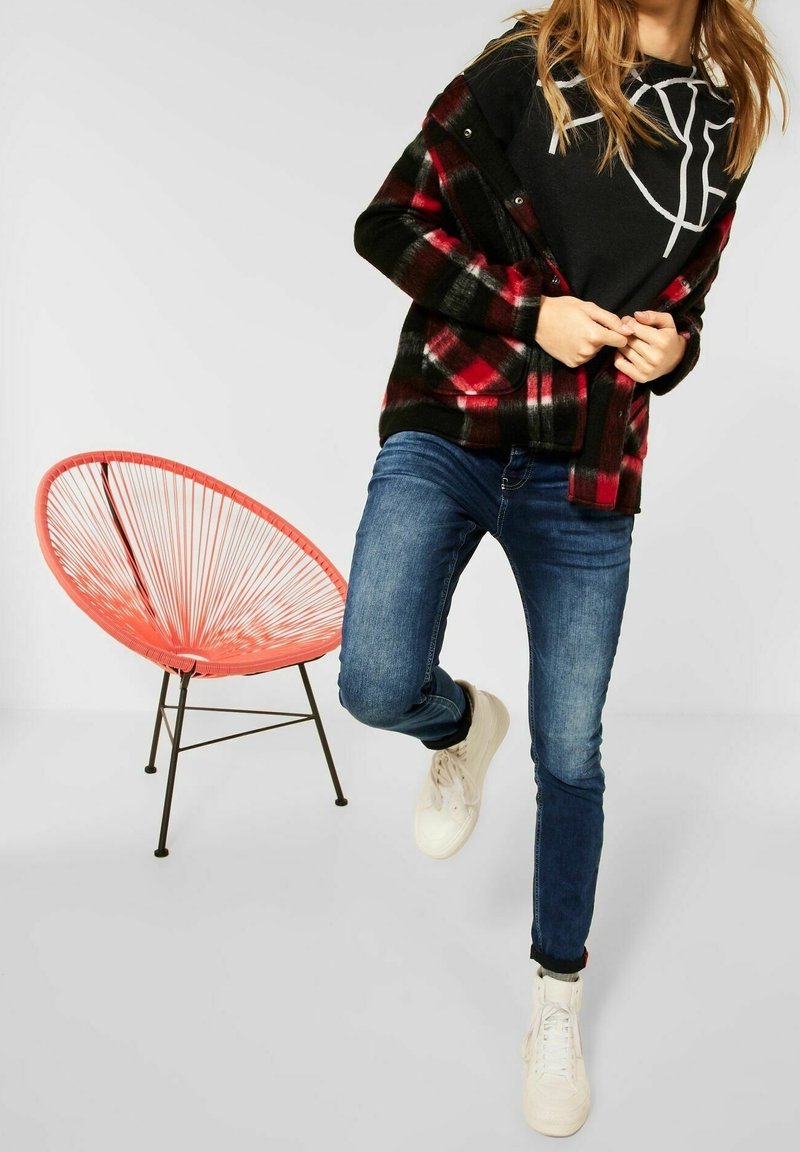 Röd och svart rutigt jacka över en svart hoodie, tillsammans med blå snäva jeans och vita sneakers. En orange stol är i bakgrunden.