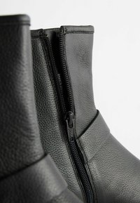 Gros plan sur des bottines en cuir noir avec des surfaces texturées et une fermeture éclair noire partiellement ouverte sur le côté.
