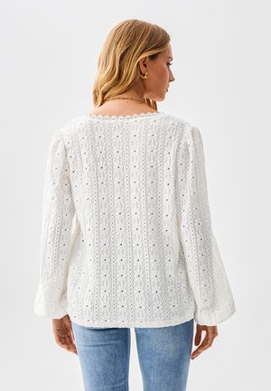 Blouse blanche brodée à manches longues, présentant un motif texturé et un décolleté festonné, portée avec un jean bleu clair.