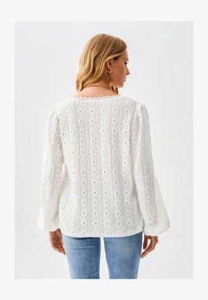 Blouse blanche brodée à manches longues, présentant un motif texturé et un décolleté festonné, portée avec un jean bleu clair.