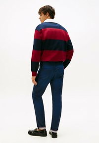 Hombre vistiendo un suéter a rayas azul marino y rojo, vaqueros oscuros con puños, y mocasines negros, de pie ante un fondo blanco liso.