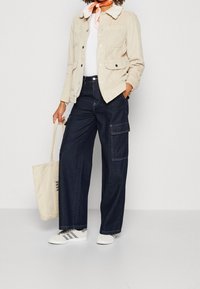 Chaqueta de pana beige con cuello de pelo, camiseta blanca, pantalones cargo oscuros de pierna ancha, zapatillas grises y una bolsa de lona con texto negro.