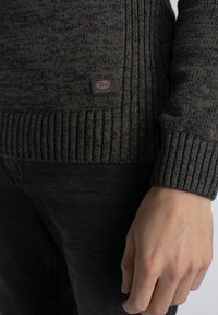 Maglione lavorato a maglia di un grigio scuro con polsini a costine e una piccola etichetta con il marchio in basso a sinistra. Texture liscia e design aderente.