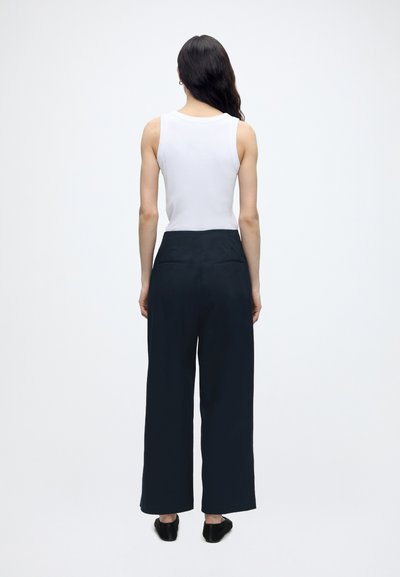 Femme debout, dos à la caméra, vêtue d'un haut blanc sans manches, d'un pantalon noir à jambes larges et de chaussures noires sur un fond blanc uni.