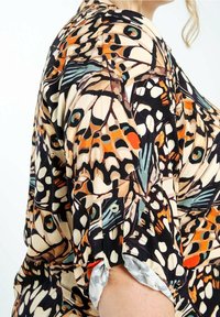 Detail van de mouw van een blouse met een levendig vlinder vleugelpatroon en een knoopsluiting, gedragen aan een arm gebogen in de elleboog tegen een effen achtergrond.