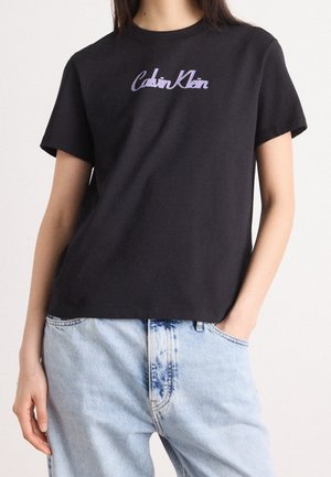 Musta Calvin Klein T-paita, jota käyttää henkilö, jolla on vaaleansiniset farkut, oikea käsi taskussa, yksivärisen valkoisen taustan edessä.