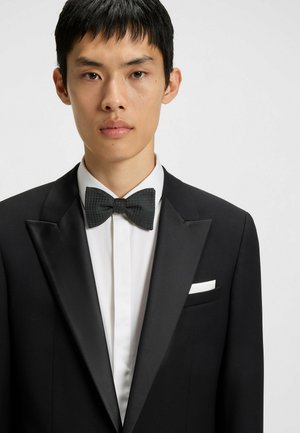Giacca da smoking nera con rever in satin, camicia bianca e cravattino nero testurizzato. Il fazzoletto da taschino è piegato ordinatamente nella tasca sinistra.