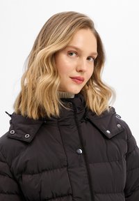 Czarna kurtka puchowa z zamkiem błyskawicznym, zatrzaskami i odpinanym kapturem. Posiada pikowany materiał i wysoki kołnierz.