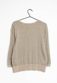 Maglione a maglia beige con pois neri, scollo rotondo, maniche lunghe e orlo a coste. Pende da un gancio nero su uno sfondo bianco.