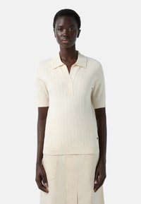 Pletený polo svetr v off-white barvě s kr�átkými rukávy, vertikálním žebrováním, véčkovým výstřihem a malým límcem. Hladká textura a přiléhavý střih.