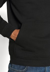 Sudadera negra de tela suave con un bolsillo frontal. Presenta puños y dobladillo acanalados, combinada con jeans de mezclilla azul.