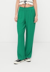 Pantalon vert à jambes larges avec un tissu texturé, une taille élastique et deux poches avant. Associé à des baskets noires.