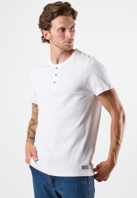 Valkoinen lyhythihainen t-paita, jossa on resoitetut pinnat, kolmipainikkeinen nappilista ja pieni logotunniste helmassa. Yhdistetty sinisiin farkkuihin.