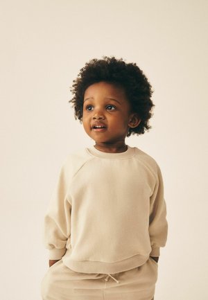 Småbarn med krøllet hår iført beige sweatshirt og bukser, der kigger let til siden med hænderne i lommerne mod en ensfarvet baggrund.