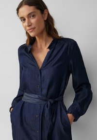 Denim shirtjurk in donkerblauw met een gerimpelde kraag, lange mouwen, gouden knopen en een strikceintuur. Gladde textuur; casual ontwerp.