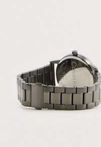 Ασημένιο εντυπωσιακό ρολόι χειρός Calvin Klein με σύνδεσμο, τοποθετημένο οριζόντια ώστε να φαίνεται η πίσω όψη και το κούμπωμα σε ανοιχτό φόντο.