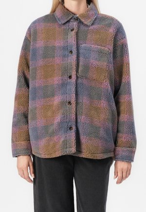 Langærmet fleece skjortejakke med brunt, gråt og pink ternet mønster, knapper foran, brystlomme, båret over sorte bukser.