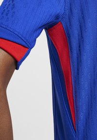 Camiseta deportiva azul con tejido texturizado, acentos rojos en las mangas y un diseño ajustado con una abertura lateral para mayor transpirabilidad.