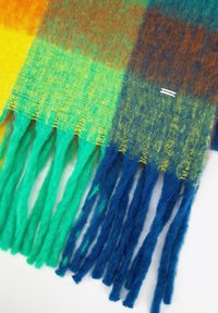 Kolorowy szalik w kratę wykonany z miękkiego, fakturowanego materiału. Zdobiony frędzlami w różnych odcieniach niebieskiego, zielonego, żółtego i brązowego.