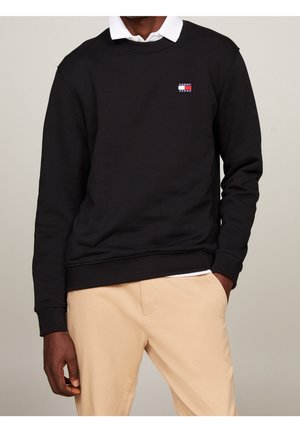 Mężczyzna w czarnej bluzie Tommy Jeans na białej koszuli z kołnierzykiem i beżowych spodniach z jedną ręką w kieszeni na jednolitym tle.