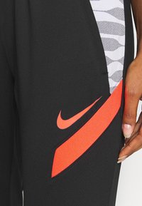 Zwarte sportbroek met een zijzak met een grijs-witte zigzagpatroon, aangevuld met een feloranje Nike-logo en accent.