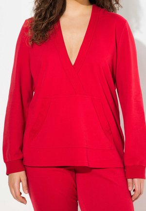 Frau trägt ein leuchtend rotes Langarm-V-Ausschnitt-Oberteil mit Fronttasche und passende rote Hose, steht vor einem schlichten Hintergrund.